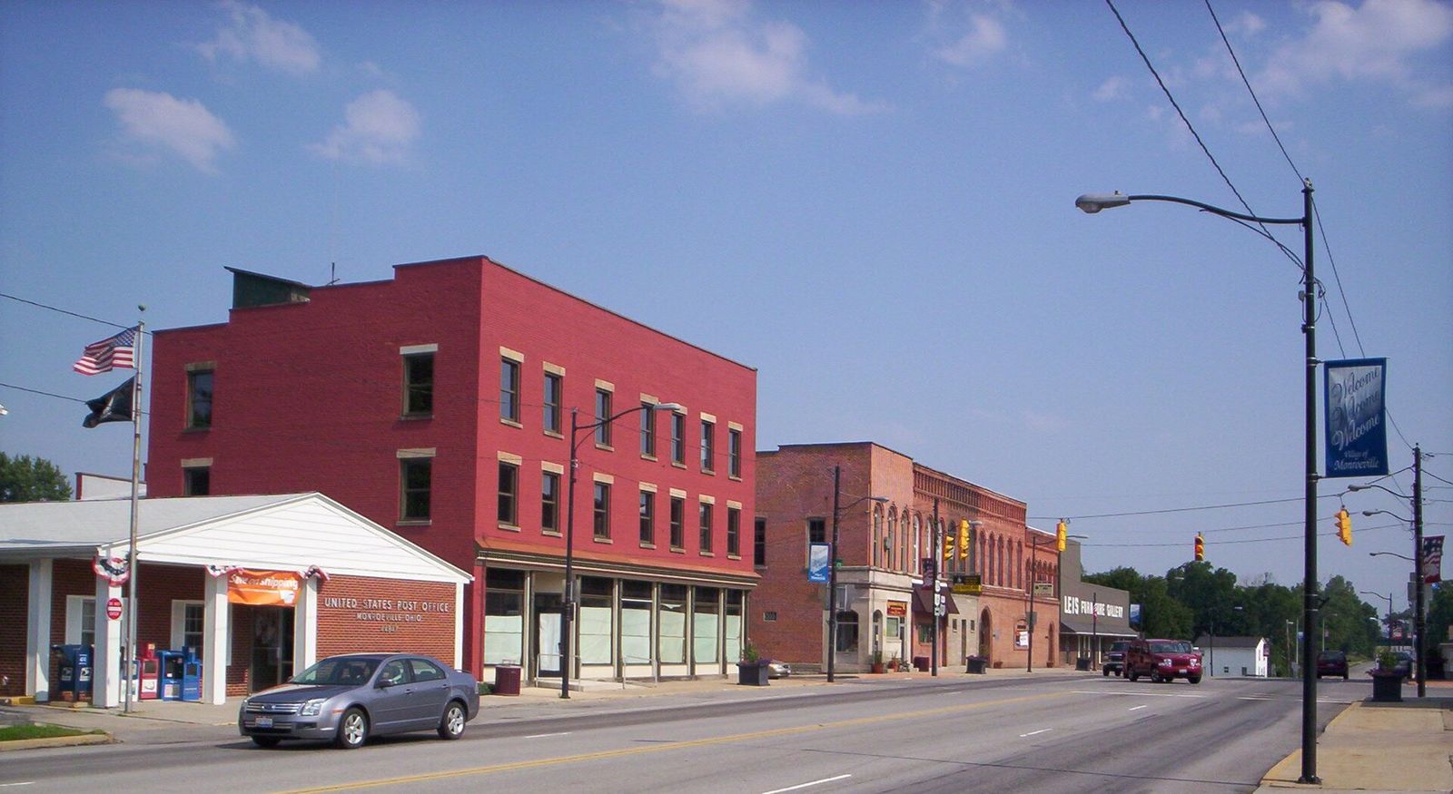 Main Street in downtown Monroeville: a three-story red brick block beside the one-story post office, with a row of brick storefronts beyond it under a blue sky. / La calle Main en el centro de Monroeville: un edificio de ladrillo rojo de tres pisos junto a la oficina de correos de un piso, con una hilera de tiendas de ladrillo detrás bajo un cielo azul.