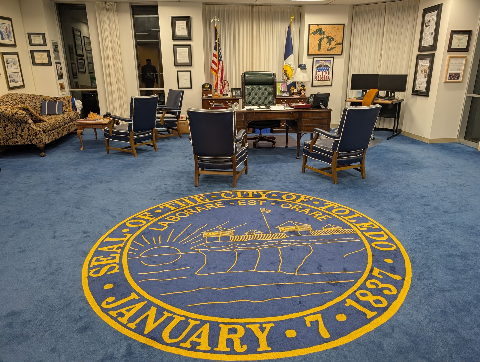 The Toledo mayor's office: the city seal, Seal of the City of Toledo, January 7, 1837, woven in gold into a blue carpet, four navy leather chairs facing a wooden desk with a green leather chair, the U.S. and Toledo flags behind it, and framed front pages and certificates on the walls. / La oficina del alcalde de Toledo: el sello de la ciudad tejido en dorado sobre una alfombra azul, cuatro sillas de cuero azul marino frente a un escritorio de madera con un sillón de cuero verde, las banderas de Estados Unidos y de Toledo detrás, y portadas y diplomas enmarcados en las paredes.