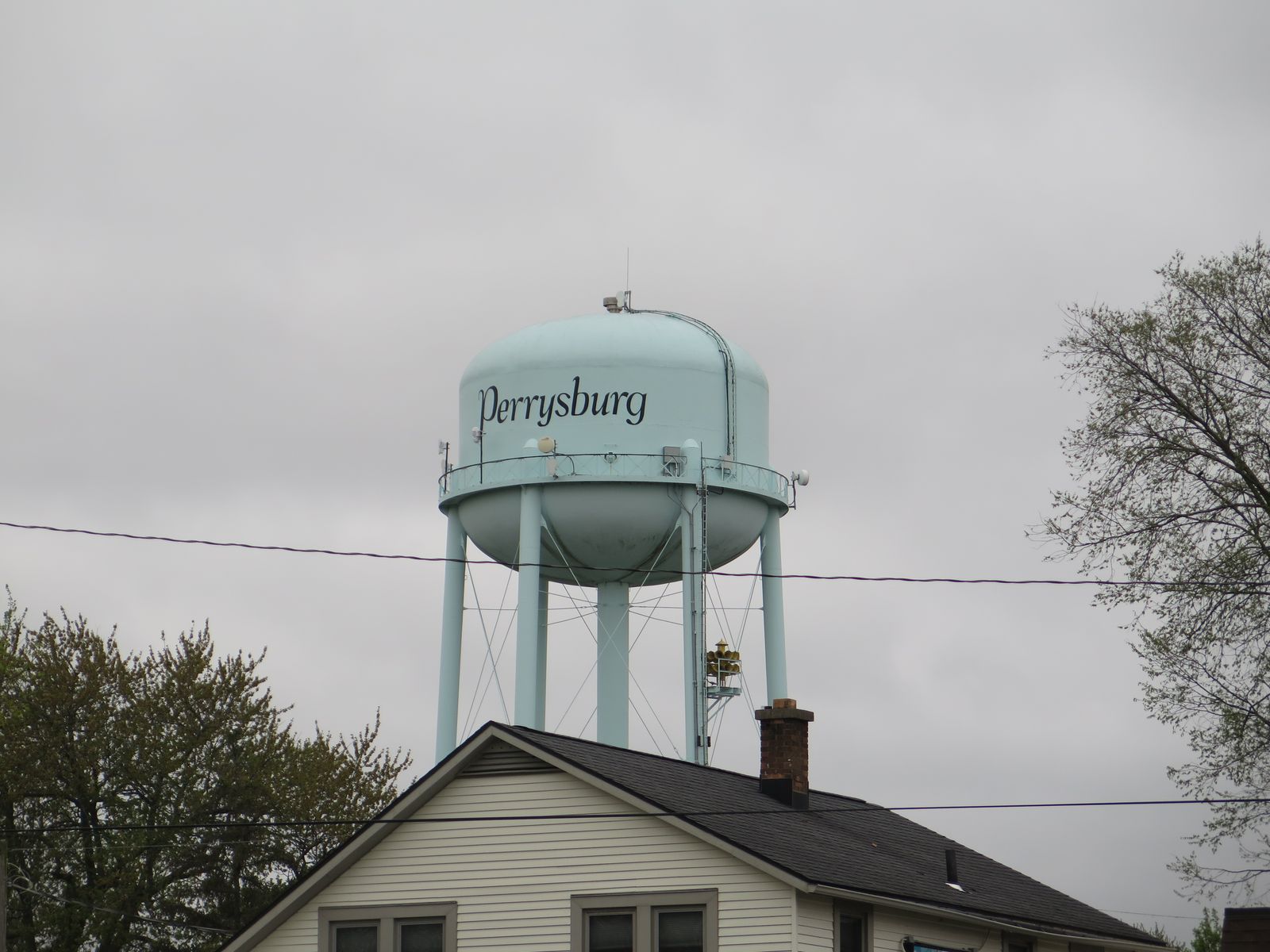 The Perrysburg, Ohio, water tower against an overcast sky: a pale blue-green tank on four legs with the word Perrysburg painted across it in black script, standing above the roof and brick chimney of a house and bare spring trees. / La torre de agua de Perrysburg, Ohio, contra un cielo nublado: un tanque azul verdoso pálido sobre cuatro patas con la palabra Perrysburg pintada en letra cursiva negra, sobre el techo y la chimenea de ladrillo de una casa y árboles primaverales sin hojas.