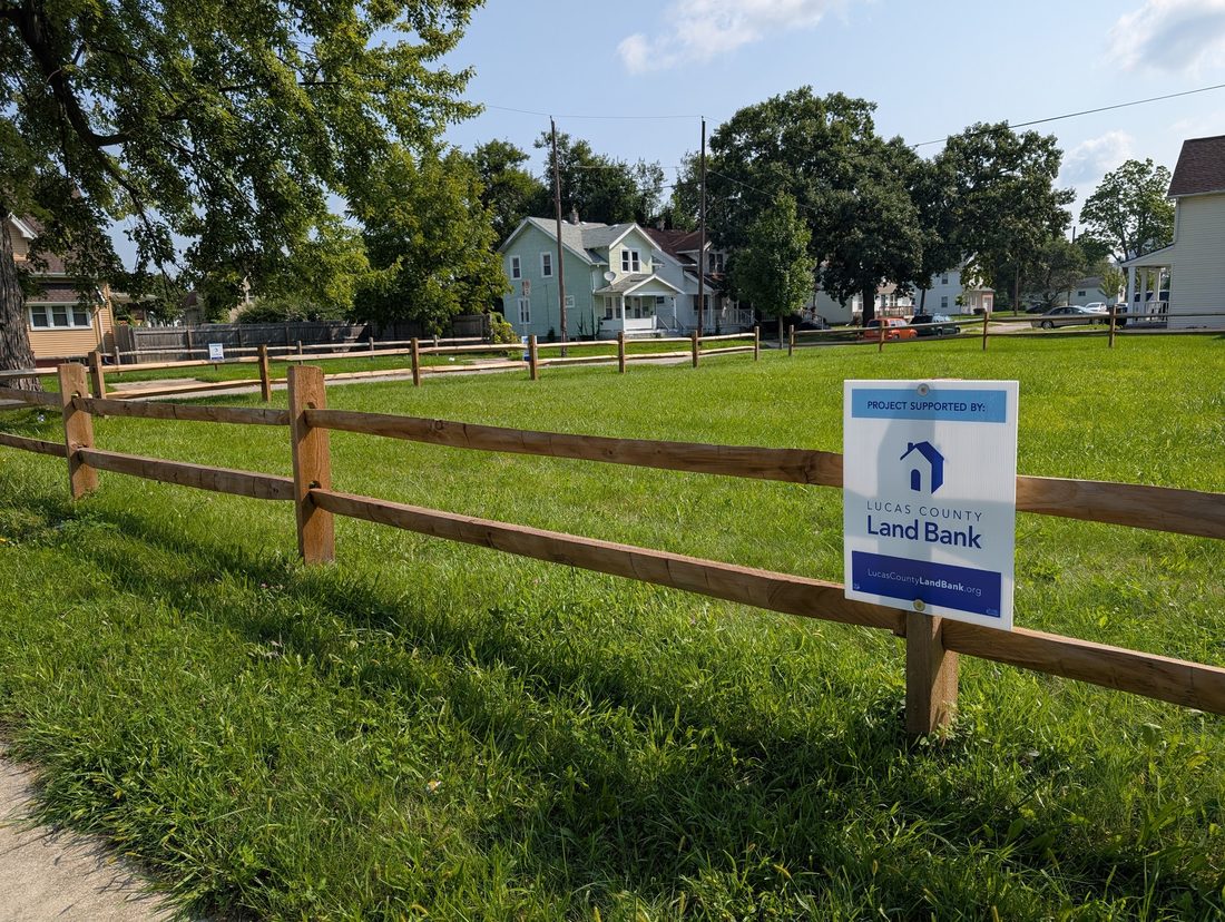 A mown vacant lot on City Park Avenue behind a new split-rail fence, with a Lucas County Land Bank sign attached to it and houses across the street.