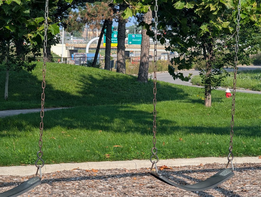 Two empty playground swings in the foreground; beyond them, through trees, green interstate signs reading I-75 North Detroit and TO 23 Sylvania Ann Arbor.