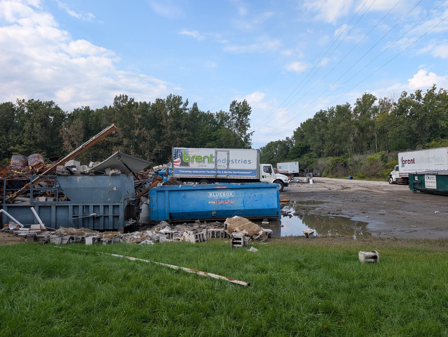 Two Brent Industries box trucks parked intact on a gravel lot beside the wreckage of the company's plant: a crushed blue-gray compactor, a blue Bluebox dumpster, shattered cinder block, rusted drums and standing water, with a treeline behind. The nearer trailer reads brent industries and, beneath it, Cleaning Industrial Textiles for a Better Environment. / Dos camiones de caja de Brent Industries estacionados intactos en un lote de grava junto a los escombros de la planta de la empresa: un compactador gris azulado aplastado, un contenedor azul de Bluebox, bloques de cemento destrozados, tambores oxidados y agua estancada, con una arboleda al fondo. El remolque más cercano dice brent industries y, debajo, Cleaning Industrial Textiles for a Better Environment.