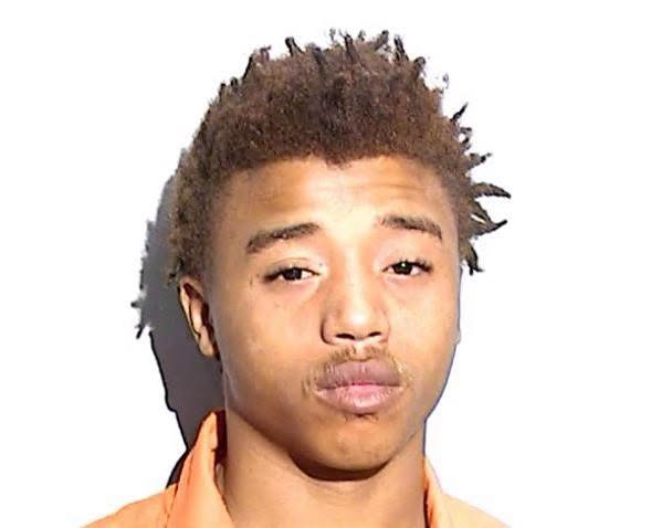 A police booking photograph of a young man in an orange jail shirt against a plain white background, facing the camera. / Una fotografía de fichaje policial de un joven con camisa naranja de la cárcel sobre un fondo blanco liso, mirando a la cámara.