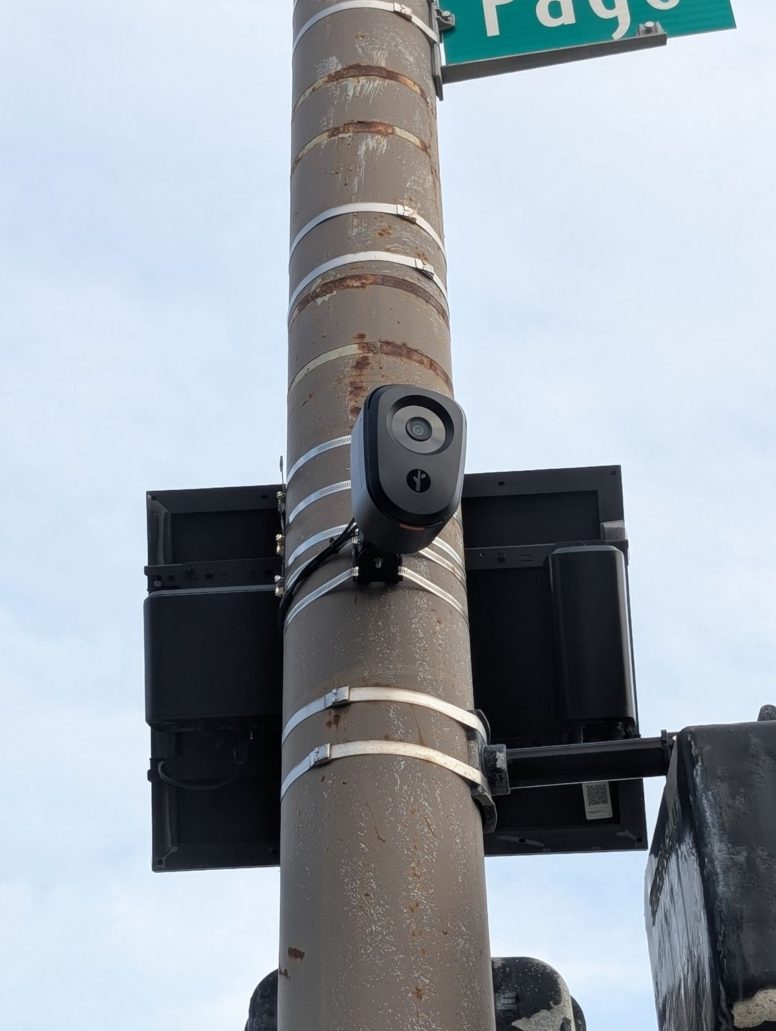 A dark, rounded Flock Safety license-plate camera banded to a city traffic-signal mast between two signal heads, with the green Page street blade above it. / Una cámara lectora de placas de Flock Safety, oscura y redondeada, sujeta con bandas a un mástil de semáforo entre dos cabezales, con el letrero verde de la calle Page encima.