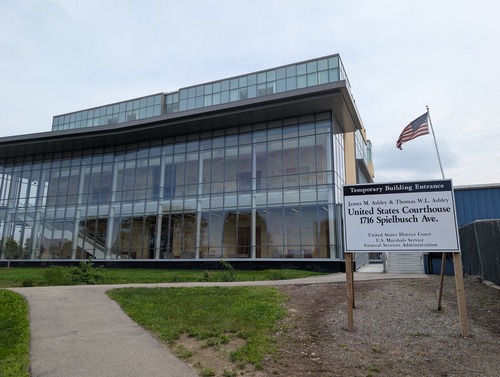 The federal courthouse in Toledo, a curtain-wall-and-stone front under the U.S. flag / El tribunal federal en Toledo, una fachada de vidrio y piedra bajo la bandera de EE. UU.