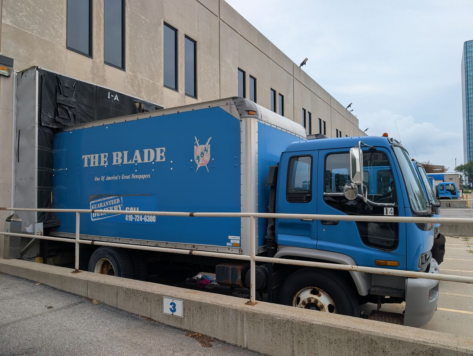 A blue delivery truck bearing The Blade’s name and a crest with crossed swords, backed up to a loading dock behind a pipe rail.