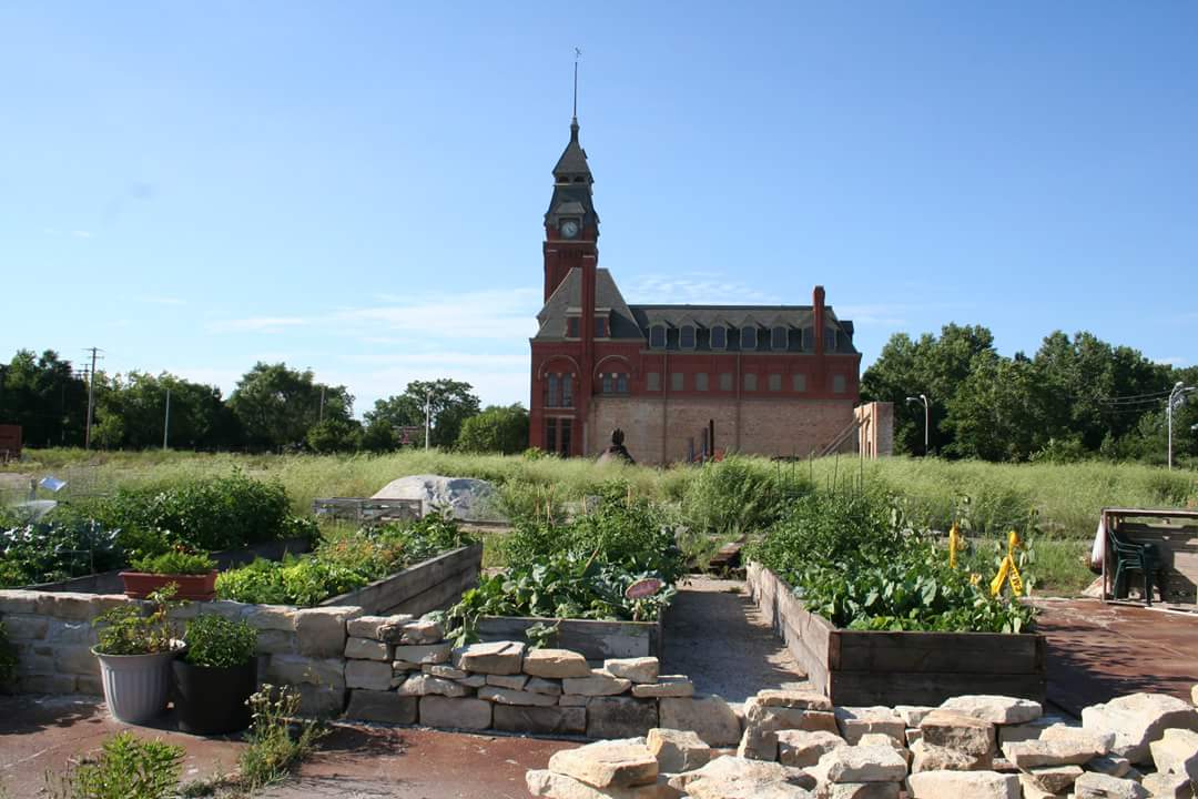 A red brick building with a tall clock tower stands behind raised vegetable beds and a low stone wall. / Un edificio de ladrillo rojo con una alta torre de reloj se levanta detrás de bancales de verduras y un muro bajo de piedra.
