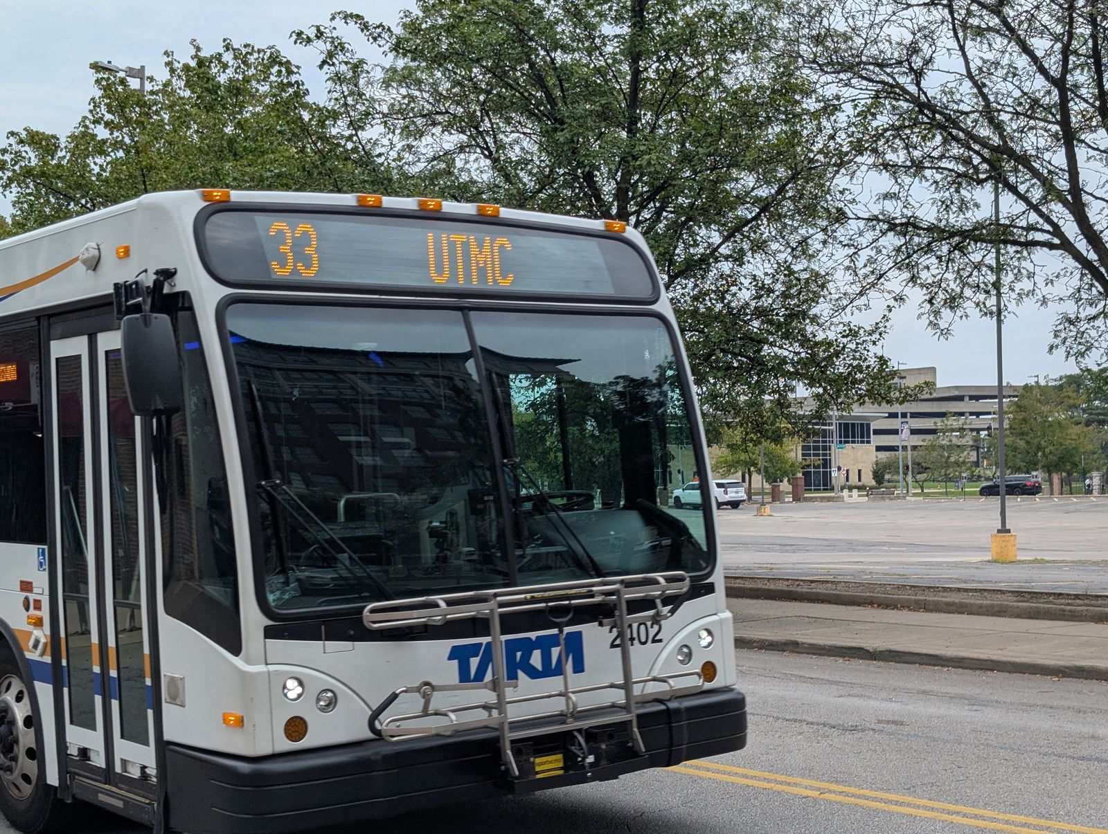 A TARTA city bus seen front three-quarter on a downtown street, its headsign reading 33 UTMC, fleet number 2402 on the bumper and a bike rack folded up at the front. / Un autobús urbano de TARTA visto de tres cuartos delanteros en una calle del centro, con el letrero 33 UTMC, el número de flota 2402 en el parachoques y un portabicicletas plegado al frente.