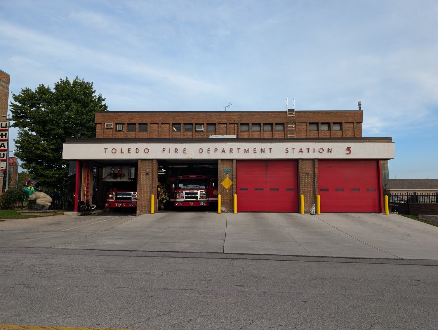 Two fire engines stand in the open bays of a brick fire house under a sign reading Toledo Fire Department Station 5. / Dos camiones de bomberos en las cocheras abiertas de una estación de ladrillo, bajo un letrero que dice Toledo Fire Department Station 5.