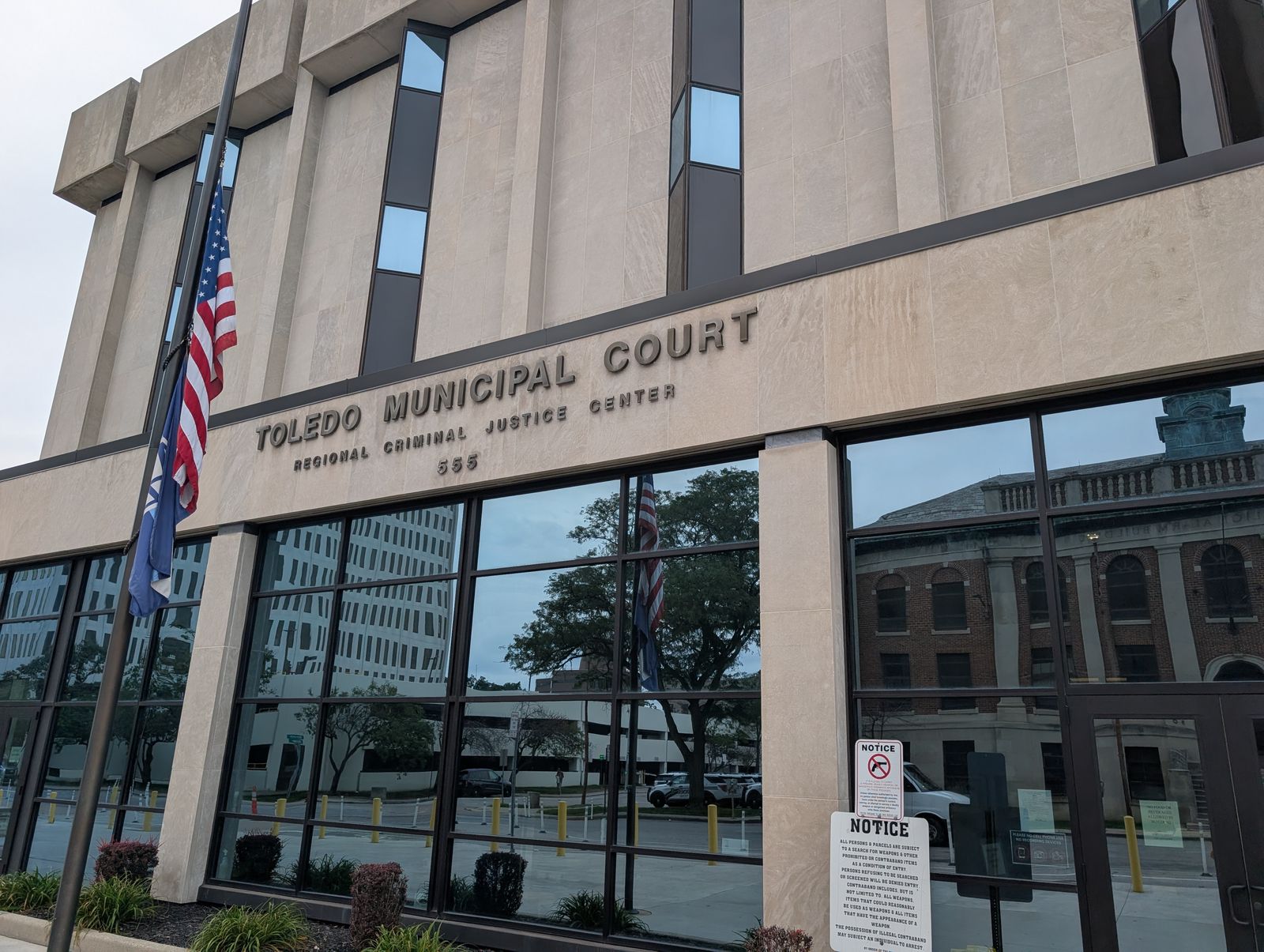 The front of the Toledo Municipal Court building, a pale stone facade lettered TOLEDO MUNICIPAL COURT, REGIONAL CRIMINAL JUSTICE CENTER, 555, with flags at the left and a notice about weapons by the glass doors. / El frente del edificio del Tribunal Municipal de Toledo, una fachada de piedra clara con las letras TOLEDO MUNICIPAL COURT, REGIONAL CRIMINAL JUSTICE CENTER, 555, con banderas a la izquierda y un aviso sobre armas junto a las puertas de vidrio.