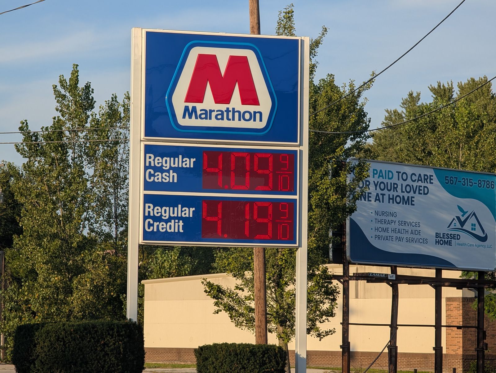 A roadside Marathon sign lit at dusk, the brand hexagon above two panels reading Regular Cash 4.09 and Regular Credit 4.19, with a utility pole, wires and a billboard behind it. / Un letrero de Marathon junto a la carretera, encendido al anochecer, con el hexágono de la marca sobre dos paneles que dicen Regular Cash 4.09 y Regular Credit 4.19, y un poste, cables y una valla publicitaria detrás.