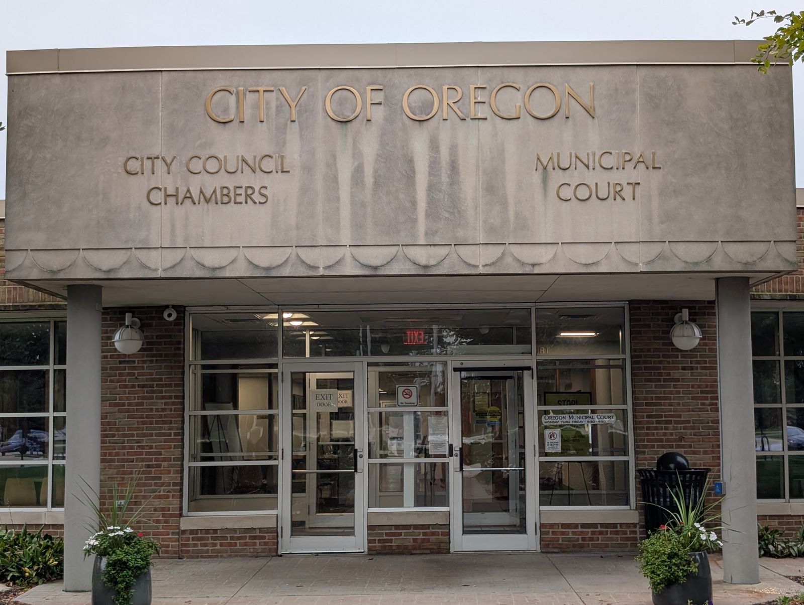 The entrance to Oregon's municipal building: CITY OF OREGON across the canopy, with CITY COUNCIL CHAMBERS and MUNICIPAL COURT lettered on either side of the glass doors. / La entrada del edificio municipal de Oregon: CITY OF OREGON en el alero, con CITY COUNCIL CHAMBERS y MUNICIPAL COURT a cada lado de las puertas de cristal.