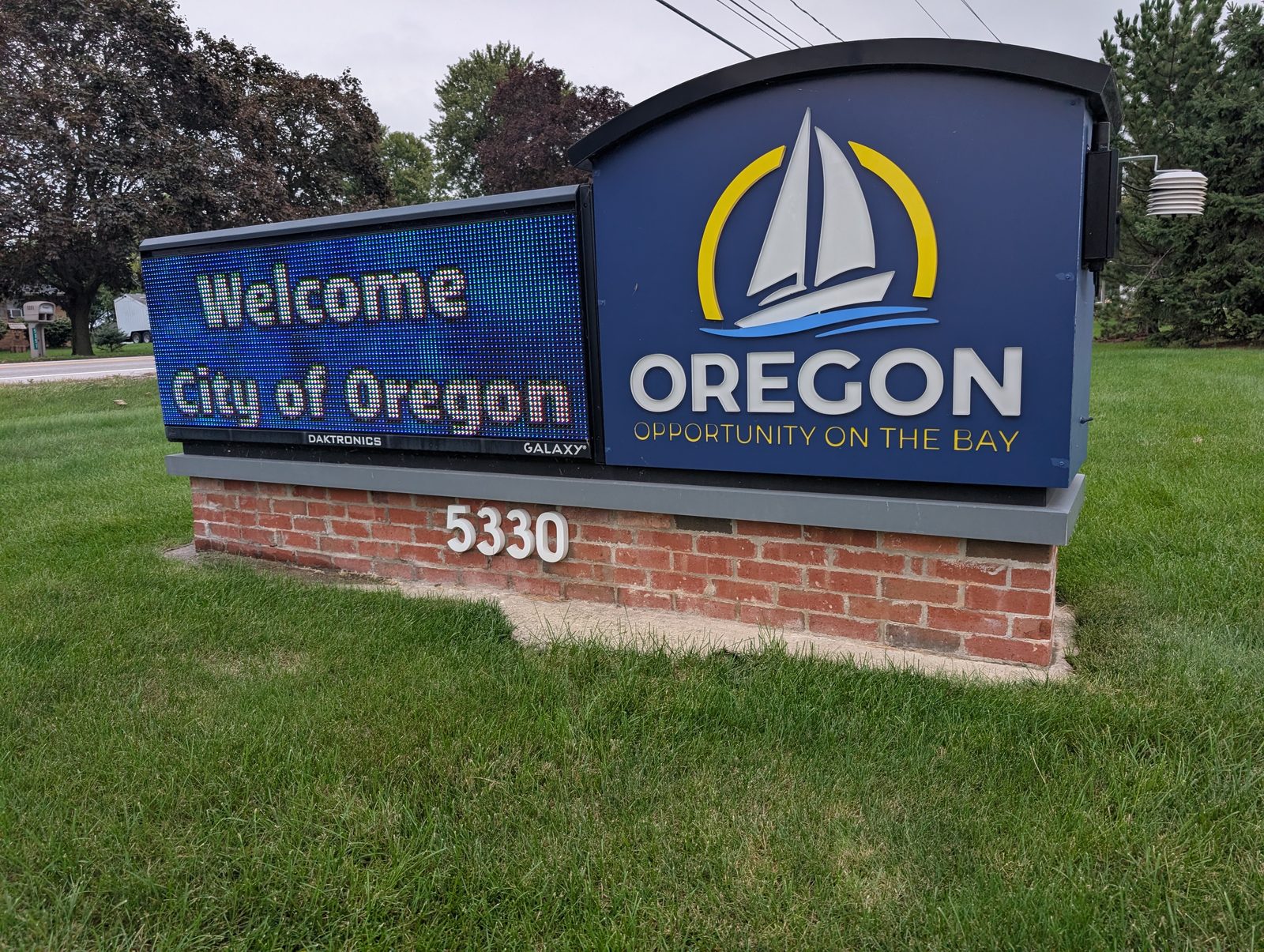 The City of Oregon’s sign on a brick base in a mowed lawn: a blue panel with a white sailboat inside a yellow arc, OREGON and OPPORTUNITY ON THE BAY, the address 5330, and an electronic message board reading Welcome, City of Oregon. Houses and trees across the road behind it, under an overcast sky. / El letrero de la Ciudad de Oregon sobre una base de ladrillo en un césped cortado: un panel azul con un velero blanco dentro de un arco amarillo, OREGON y OPPORTUNITY ON THE BAY, el número 5330 y un tablero electrónico que dice Welcome, City of Oregon. Al otro lado de la calle, casas y árboles bajo un cielo nublado.