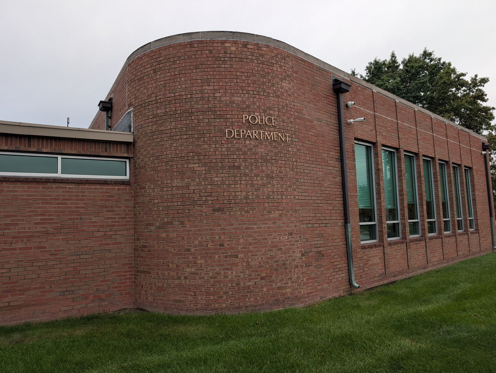 The curved red-brick corner of the Oregon police building, with POLICE DEPARTMENT in raised letters on the wall, two small security cameras mounted beside a downspout, a row of tall narrow windows and a mowed lawn in front, under an overcast sky. / La esquina curva de ladrillo rojo del edificio de la policía de Oregon, con POLICE DEPARTMENT en letras en relieve, dos pequeñas cámaras de seguridad junto a un bajante, una fila de ventanas altas y estrechas y césped cortado al frente, bajo un cielo nublado.