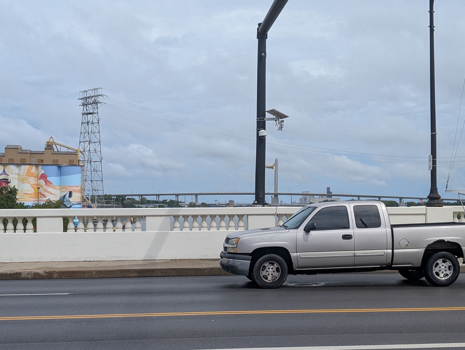 A pole-mounted camera rig over a bridge roadway — a solar panel, a plate-reader unit and a dome camera on a black pole — with a pickup truck passing beneath it and the Veterans’ Glass City Skyway downstream behind it. At the left edge, a mural on riverside grain silos and a transmission tower. / Un equipo de cámaras en un poste sobre la calzada de un puente — un panel solar, un lector de matrículas y una cámara domo en un poste negro — con una camioneta pasando debajo y el Veterans’ Glass City Skyway río abajo al fondo. En el borde izquierdo, un mural sobre silos de grano junto al río y una torre de transmisión.