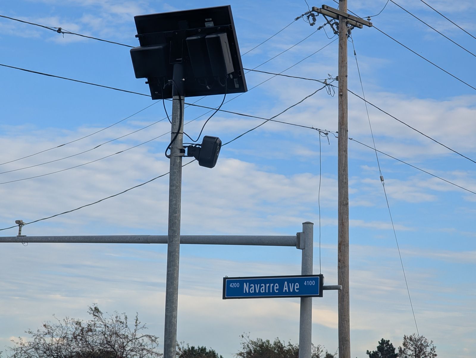 A black license plate camera clamped to a metal pole under a tilted solar panel, power lines behind it and a blue Navarre Ave street sign below, against a blue sky. / Una cámara lectora de placas negra sujeta a un poste de metal bajo un panel solar inclinado, con cables eléctricos detrás y un letrero azul de Navarre Ave debajo, contra un cielo azul.