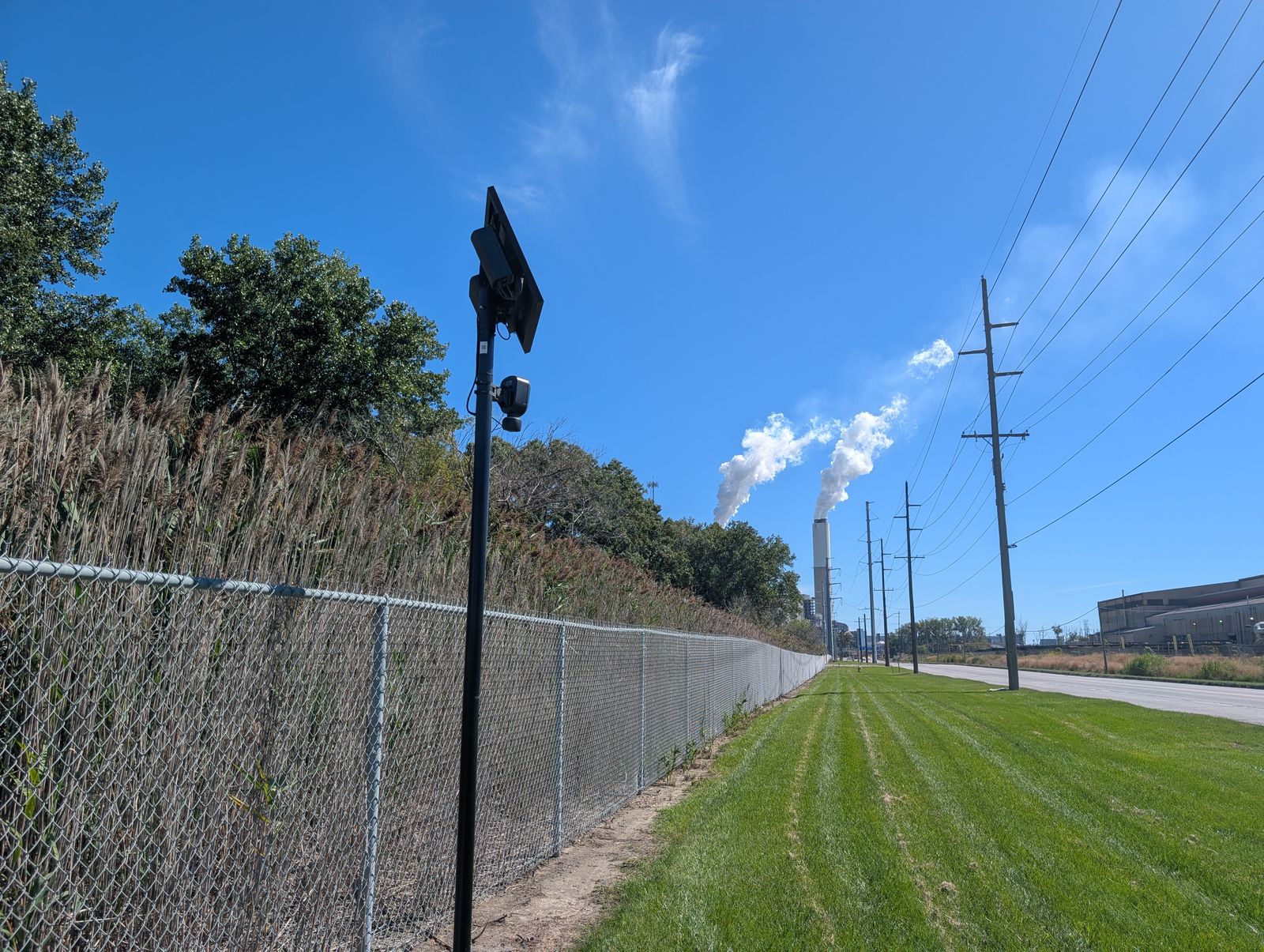A solar-powered pole carrying a Flock Safety license plate reader beside a chain-link fence and tall reeds, with a mowed strip, a road, a line of utility poles and a power plant’s smokestacks behind it. / Un poste con panel solar que sostiene una cámara lectora de placas de Flock Safety junto a una cerca de malla y cañas altas, con una franja de césped, una calle, una hilera de postes eléctricos y las chimeneas de una planta eléctrica al fondo.