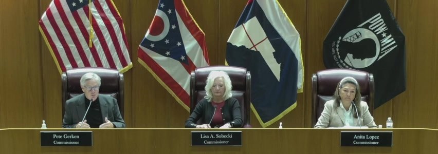 Lucas County Commissioners Pete Gerken, Lisa A. Sobecki and Anita Lopez at the dais, flags behind them, Gerken speaking into his microphone / Los comisionados del Condado de Lucas Pete Gerken, Lisa A. Sobecki y Anita Lopez en el estrado, con banderas detrás, mientras Gerken habla por su micrófono