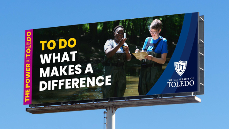 A University of Toledo billboard against a blue sky. Bold type reads TO DO WHAT MAKES A DIFFERENCE, with TO DO in gold; two students in waders take a water sample in a creek; a magenta bar up the left side reads THE POWER UTOLEDO; the university shield sits at right. / Una valla publicitaria de la Universidad de Toledo contra un cielo azul. Letras gruesas dicen TO DO WHAT MAKES A DIFFERENCE, con TO DO en dorado; dos estudiantes con botas de vadeo toman una muestra de agua en un arroyo; una franja magenta a la izquierda dice THE POWER UTOLEDO; el escudo de la universidad está a la derecha.
