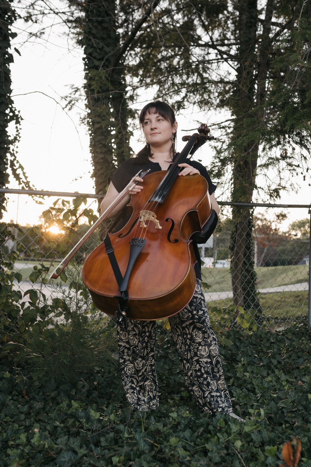 Colleen d’Alelio holds a cello and bow in front of a chain-link fence and trees. / Colleen d’Alelio sostiene un violonchelo y su arco frente a una cerca de alambre y árboles.
