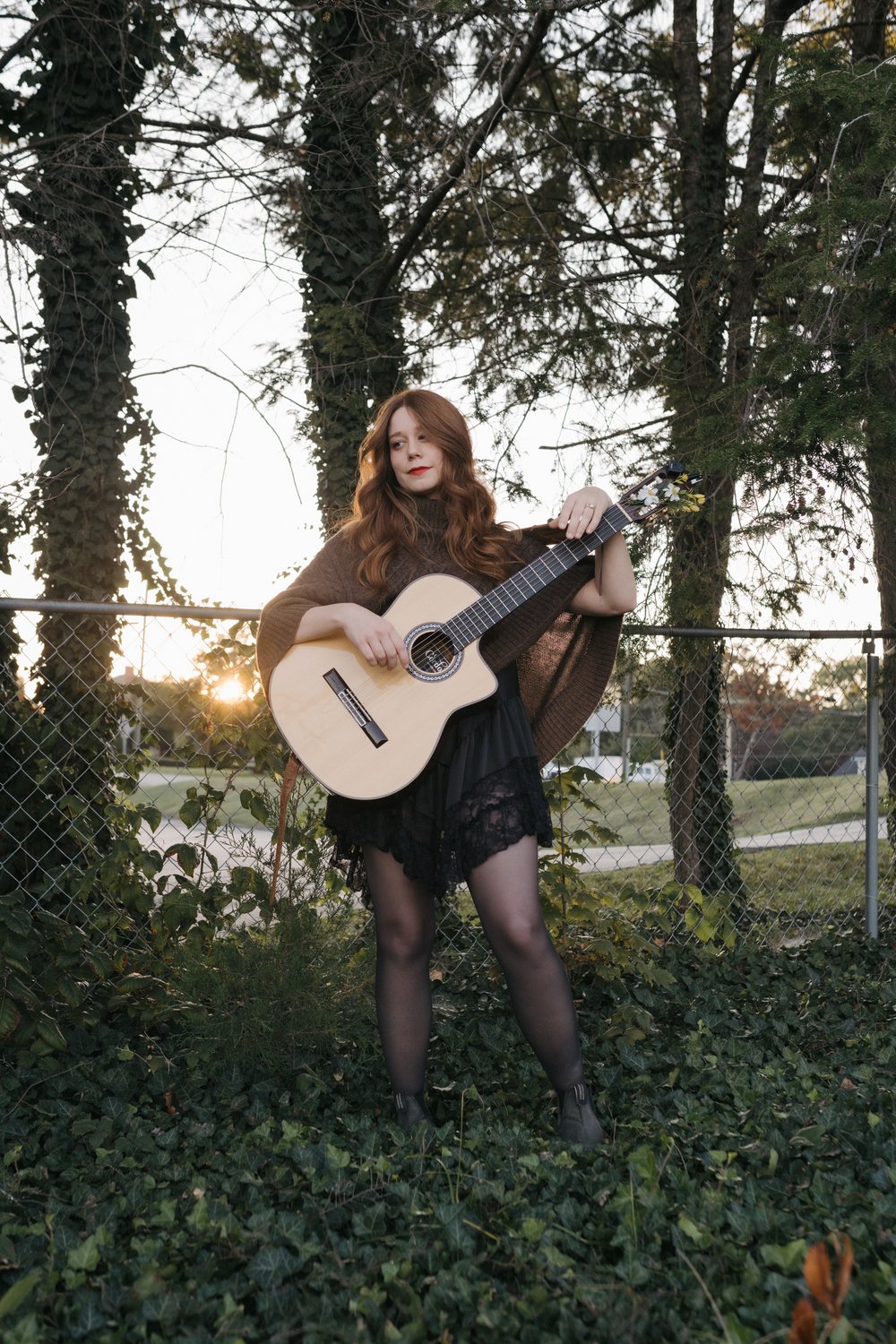 Redd Daugherty holds a guitar in front of a chain-link fence and trees. / Redd Daugherty sostiene una guitarra frente a una cerca de alambre y árboles.