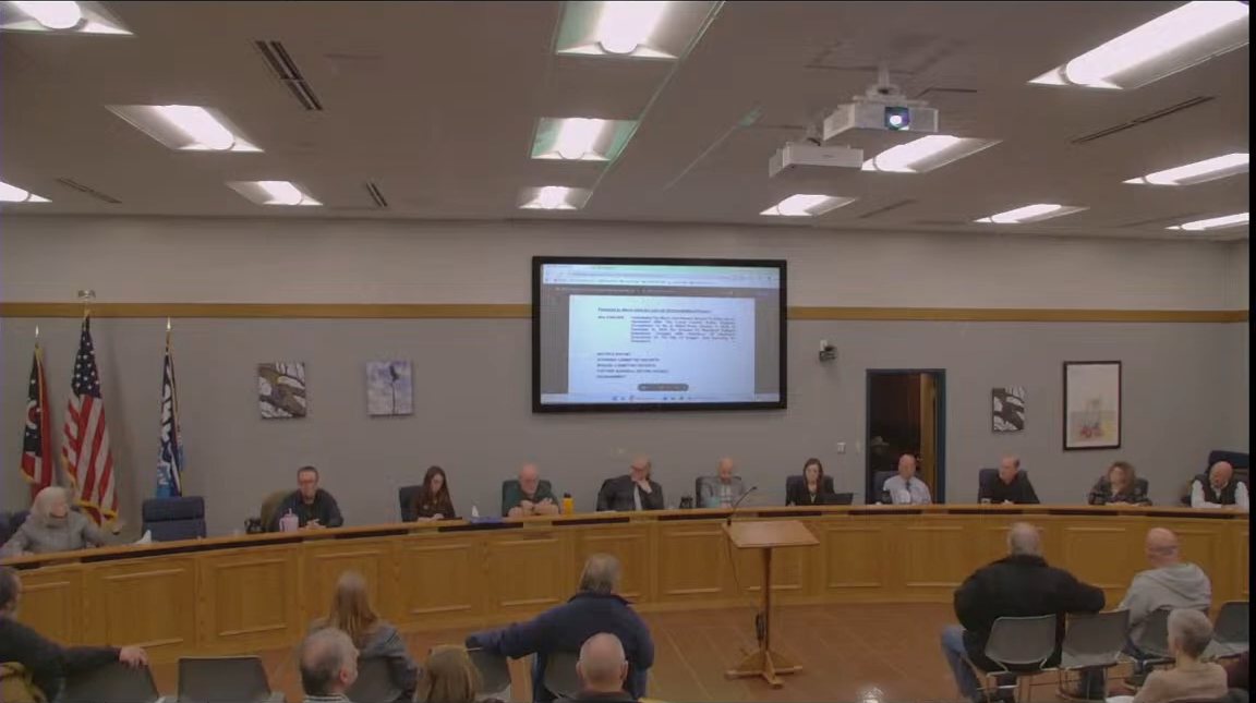 Oregon City Council members seated along a curved wooden dais under a projection screen, with flags at left and residents in folding chairs in the foreground, in the council chambers. / Miembros del Concejo Municipal de Oregon sentados en un estrado curvo de madera bajo una pantalla, con banderas a la izquierda y vecinos en sillas plegables en primer plano, en la sala del concejo.