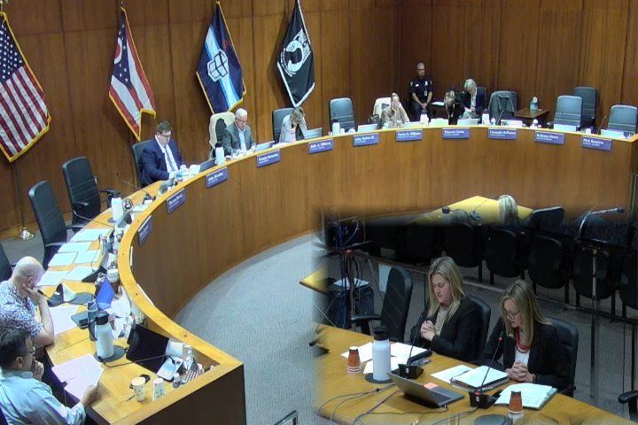 Toledo City Council members at the curved dais of council chambers, flags behind them, with an inset of two women at the presenters’ table reading from papers. / Miembros del Concejo de Toledo en el estrado curvo de la sala del Concejo, con banderas detrás, y un recuadro con dos mujeres en la mesa de los ponentes leyendo documentos.