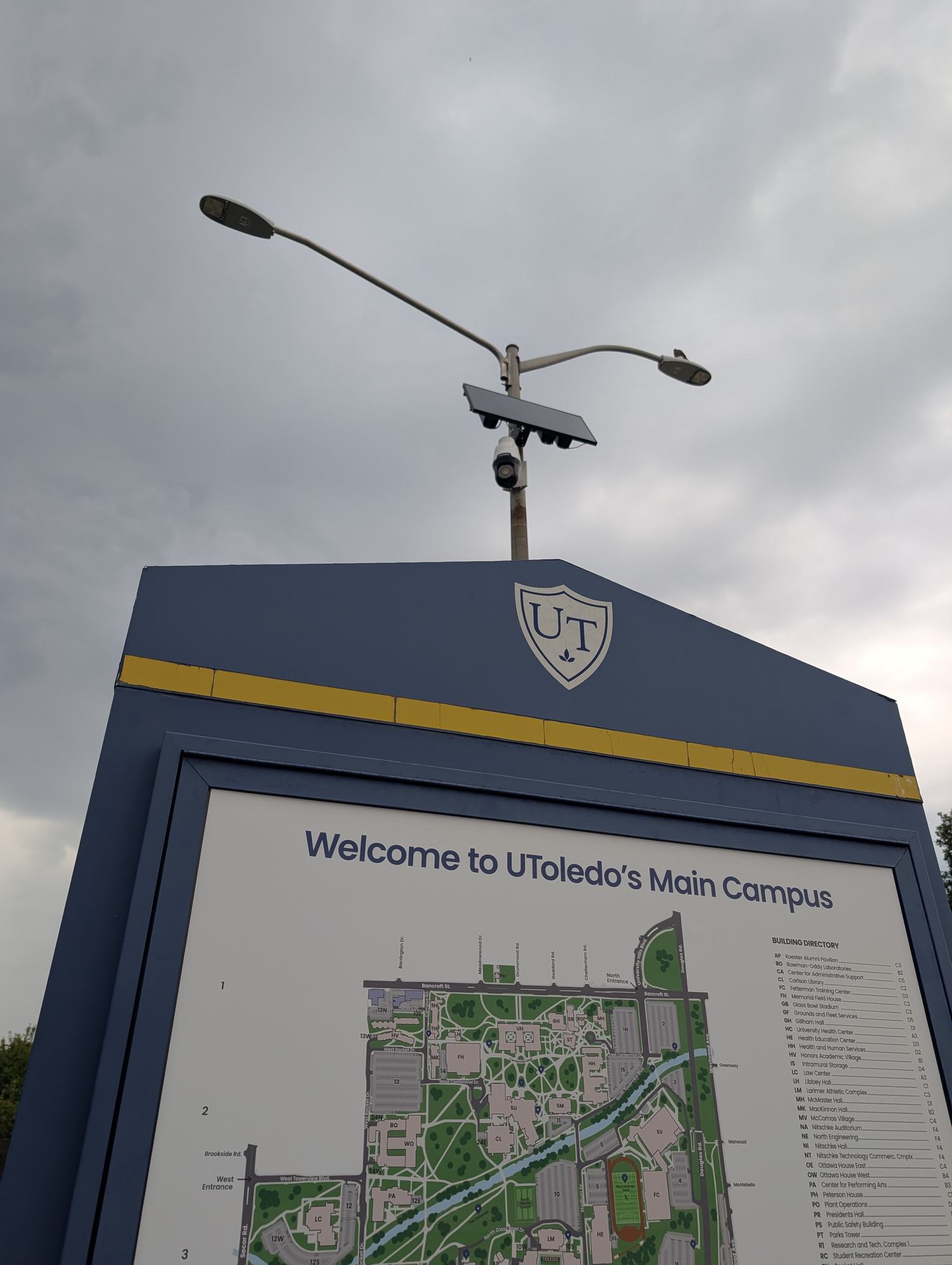 A camera pole standing directly behind and above a low brick sign reading Welcome to UToledo’s Main Campus. / Un poste con cámara que se alza justo detrás y por encima de un letrero bajo de ladrillo que dice Welcome to UToledo’s Main Campus.