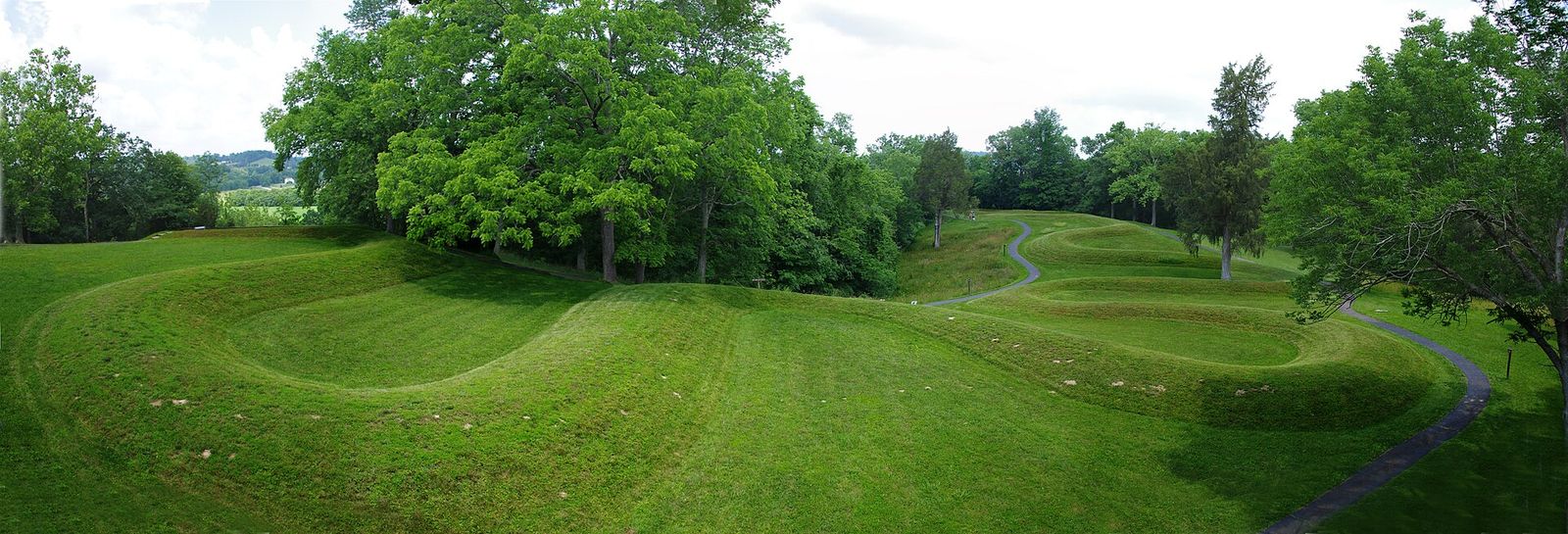 A wide view from above of the Great Serpent Mound: low grass-covered coils winding across a mown plateau, a paved footpath running beside them, trees behind.