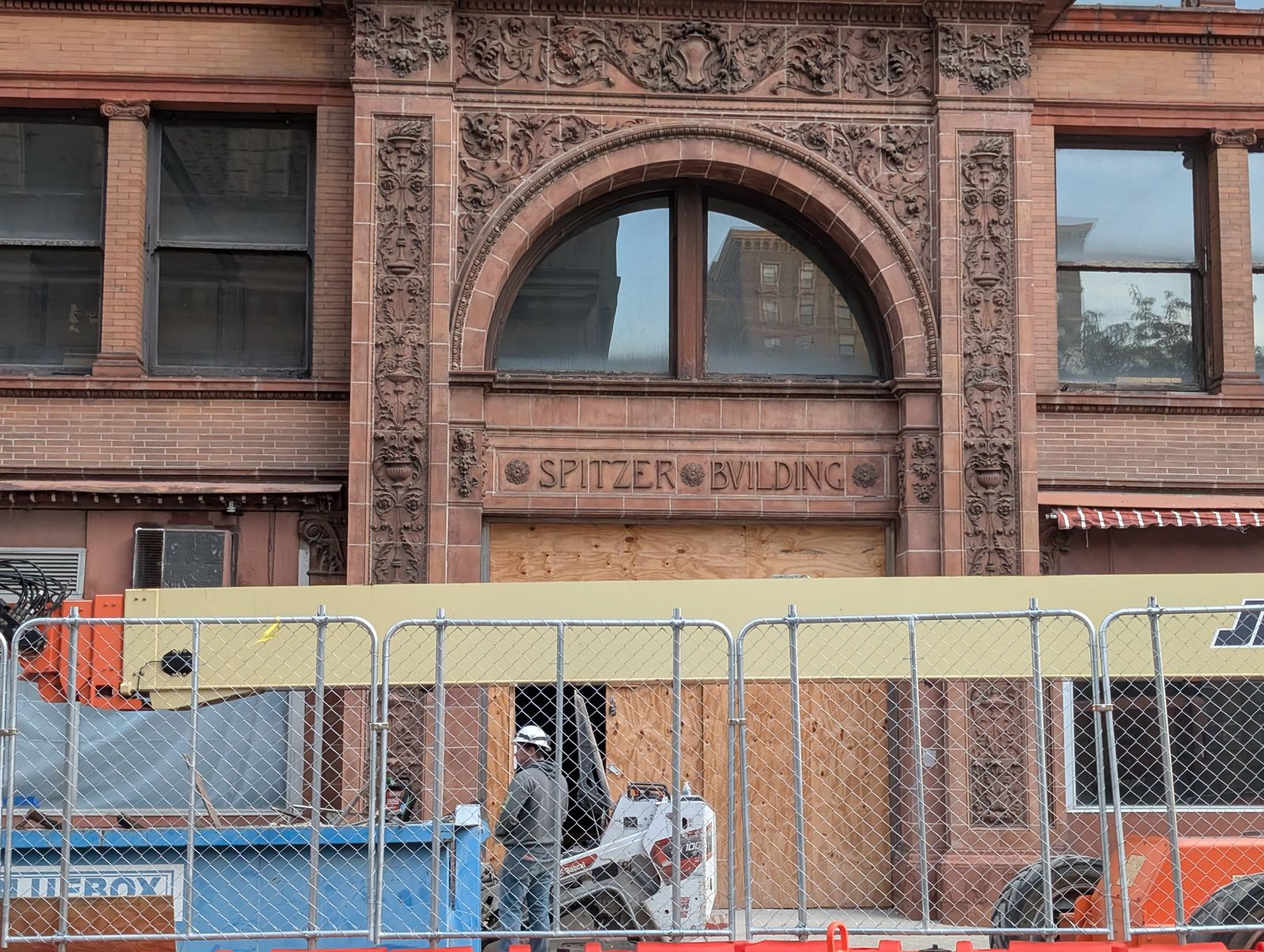 The Spitzer Building’s ornate terracotta entrance bay, its name carved into the stone above a plywood-boarded doorway, with a worker in a hard hat and a small loader behind a chain-link construction fence / La entrada de terracota labrada del Edificio Spitzer, con su nombre tallado en la piedra sobre una puerta tapiada con madera, y un obrero con casco y una pequeña cargadora detrás de una valla de obra