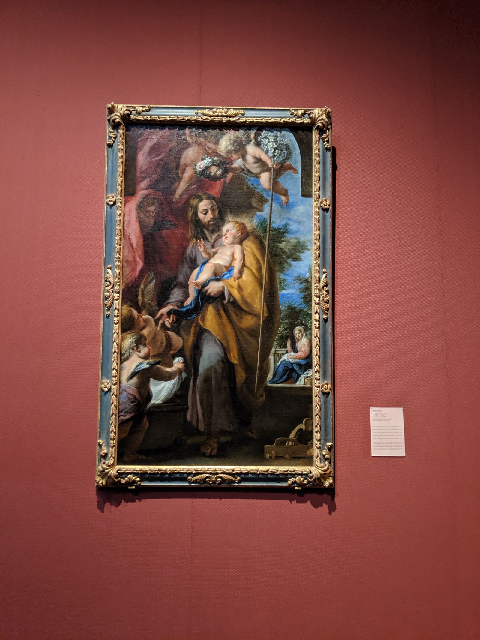 A large Baroque painting in a carved gilt frame on a dark red gallery wall: a young bearded Joseph in a yellow cloak holds the infant Christ, angels above carry a wreath of flowers, and Mary sits sewing at the right. / Un cuadro barroco grande en un marco dorado tallado, sobre una pared roja de galería: un José joven y barbado, con manto amarillo, sostiene al niño Jesús; arriba unos ángeles llevan una corona de flores y, a la derecha, María cose sentada.