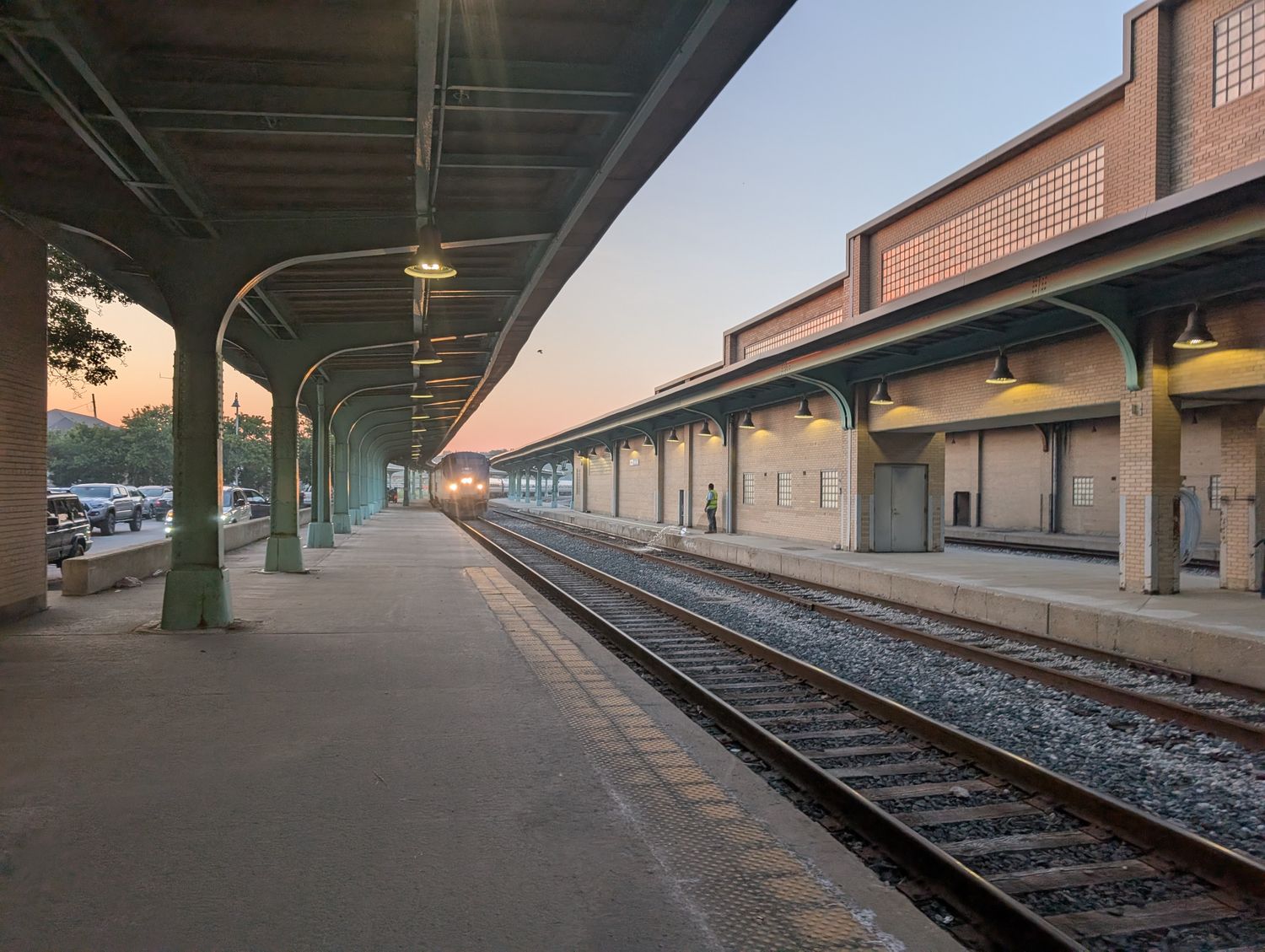 The platform at Toledo’s Amtrak station at sunrise, canopy lamps still lit, the sky going orange down the tracks. / El andén de la estación de Amtrak de Toledo al amanecer, con las lámparas de la marquesina todavía encendidas y el cielo anaranjado sobre las vías.